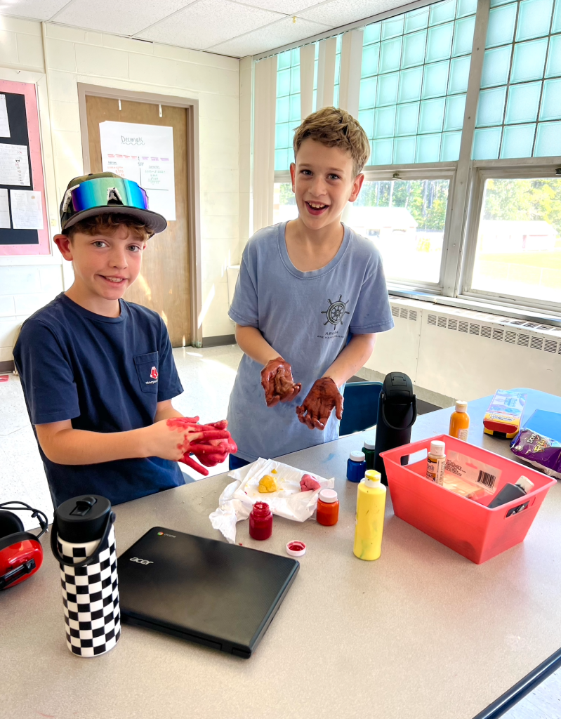 Two children with colorful paint on their hands, smiling and standing in a classroom with art supplies on the table.